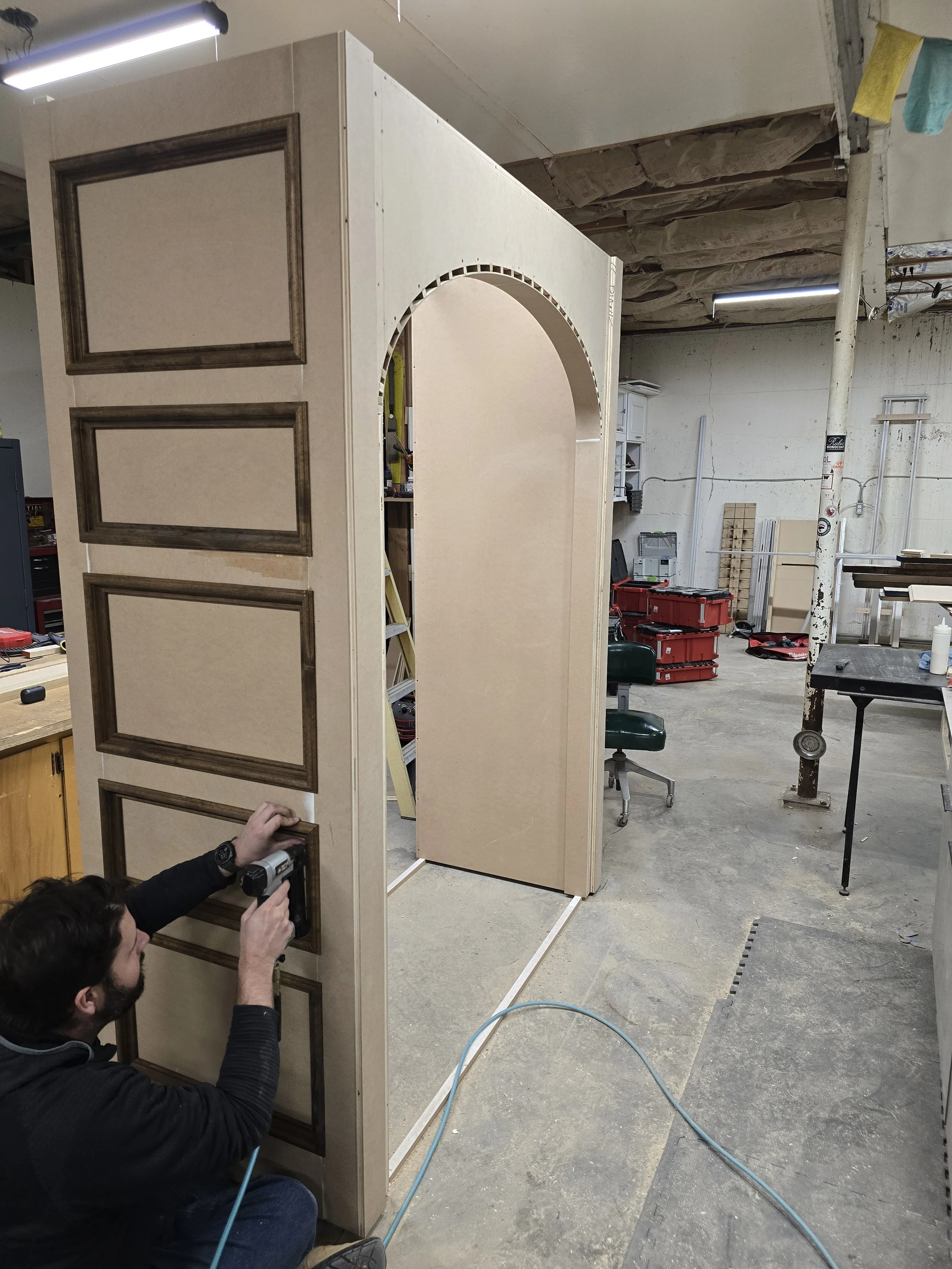 A man working on a wooden structure with painted panels in a workshop, using a cordless drill. The structure has four painted panels with decorative borders on the left side and an open archway on the right side.