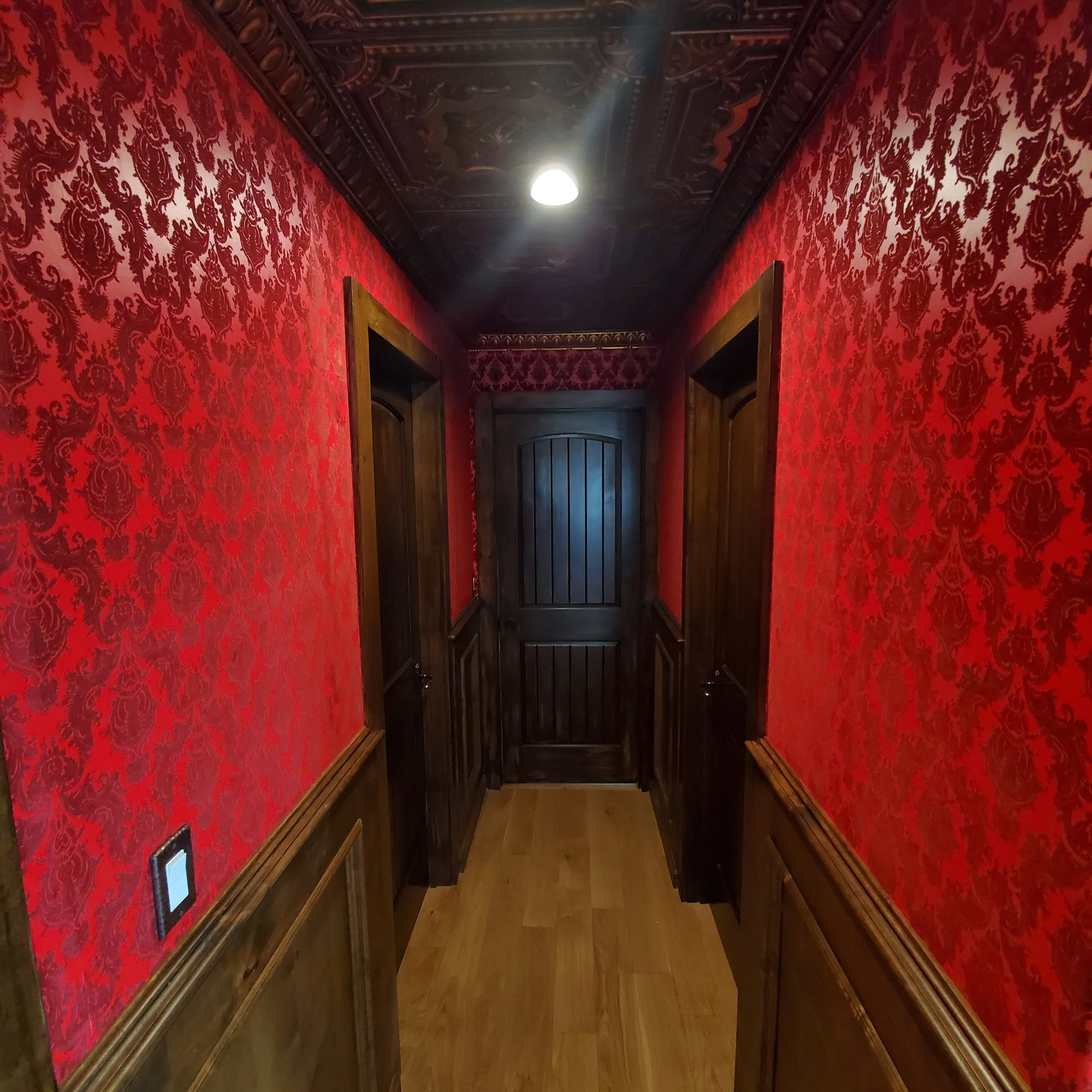 A narrow hallway with red damask wallpaper, dark wood paneling, and wooden doors on both sides, leading to a solid dark wood door at the end. The ceiling is decorated with ornate molding and has a single ceiling light.
