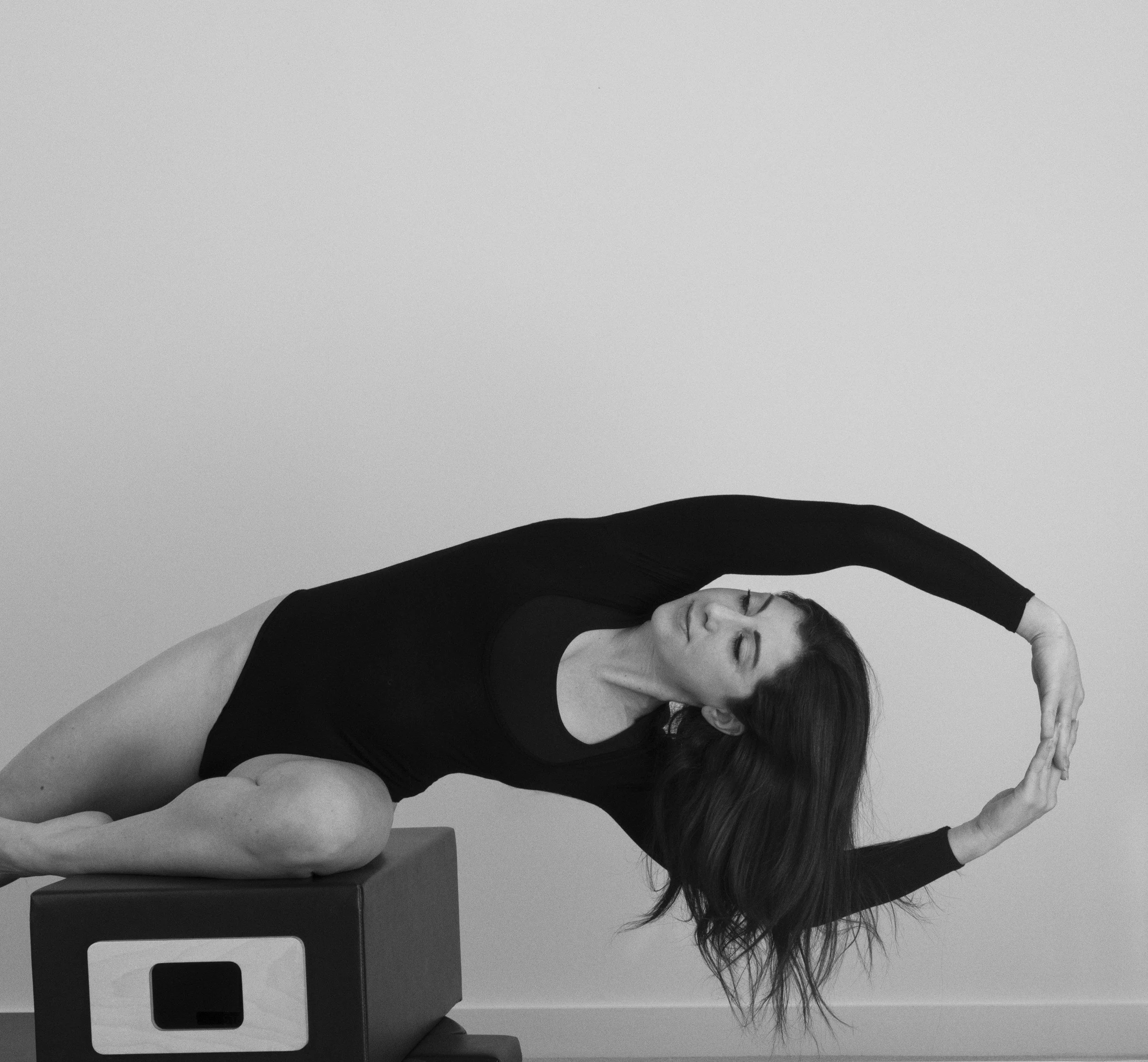 A woman with long hair, dressed in black, practicing yoga or stretching on a table in a minimalistic room with plain light-colored walls.