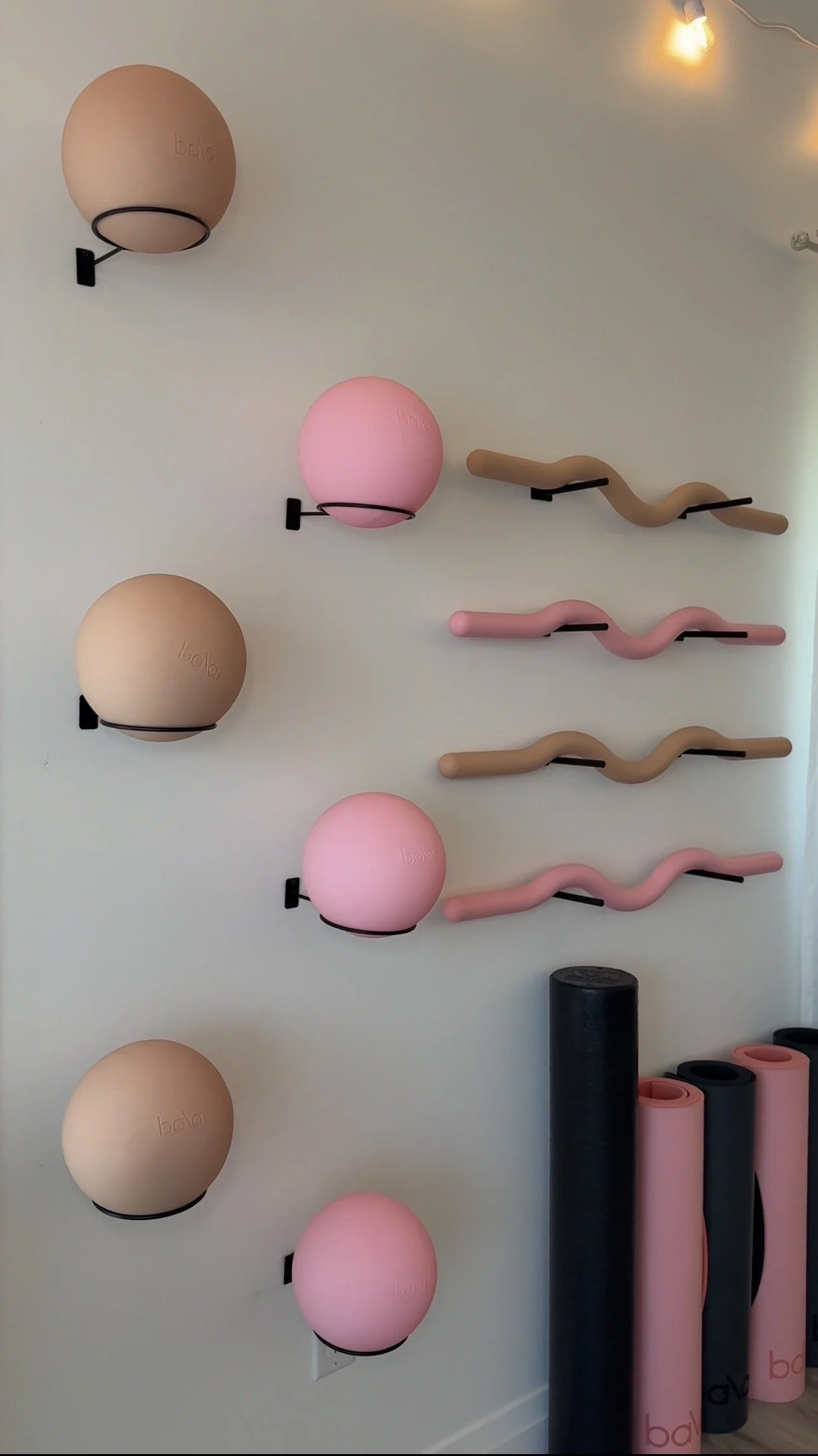 Pink and beige exercise balls mounted on a white wall with black wall mounts. Some black, pink, and gray foam rollers are on the floor below the wall.