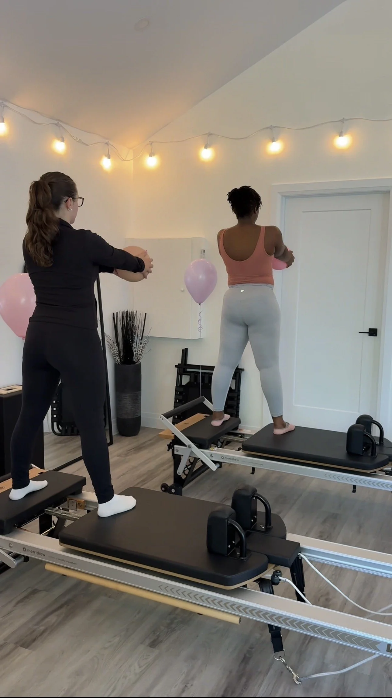 A woman is standing on a Pilates reformer machine, performing an exercise with her arms crossed, while a trainer assists her. The room has string lights on the ceiling, a balloon, and some decorative plants.
