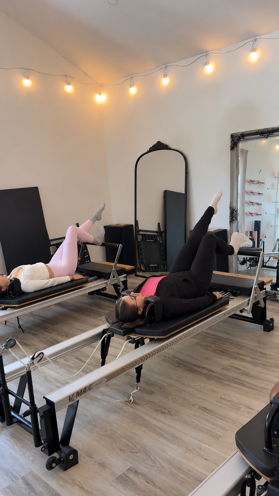 Two women exercising on Pilates reformers in a fitness studio with string lights and a mirror.