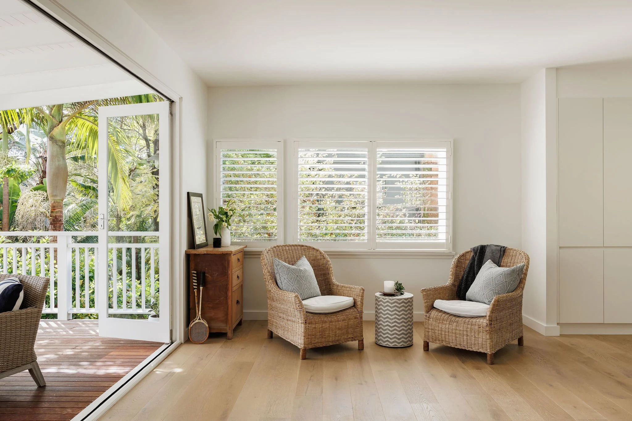 A bright living room with wicker chairs, a small round table, a wooden cabinet, and large windows with white shutters, opening to a balcony with trees outside.