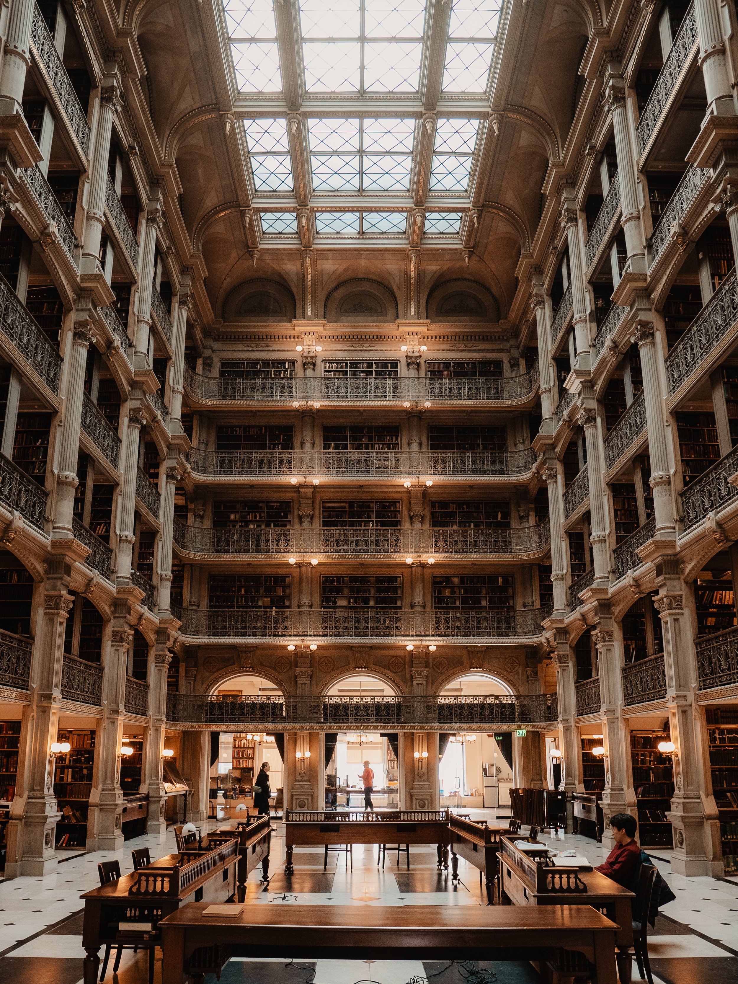 Interior of a grand historic library with multiple floors of bookshelves, ornate railings, large arched windows, and a glass ceiling allowing natural light. Several people are reading or walking inside.