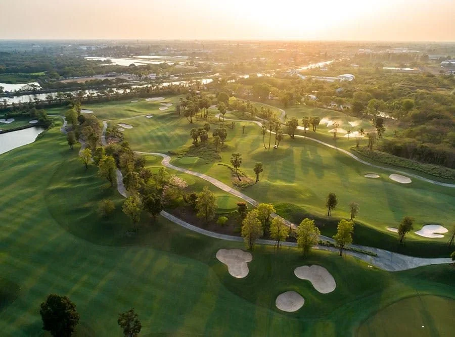 Aerial view of a golf course at sunset, featuring green fairways, sand traps, trees, and water hazards.
