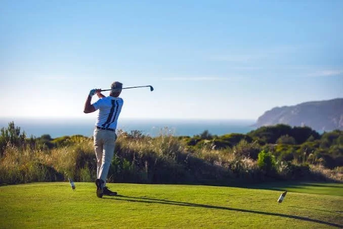 A person playing golf on a green course near the coast, swinging a club as the sun shines.