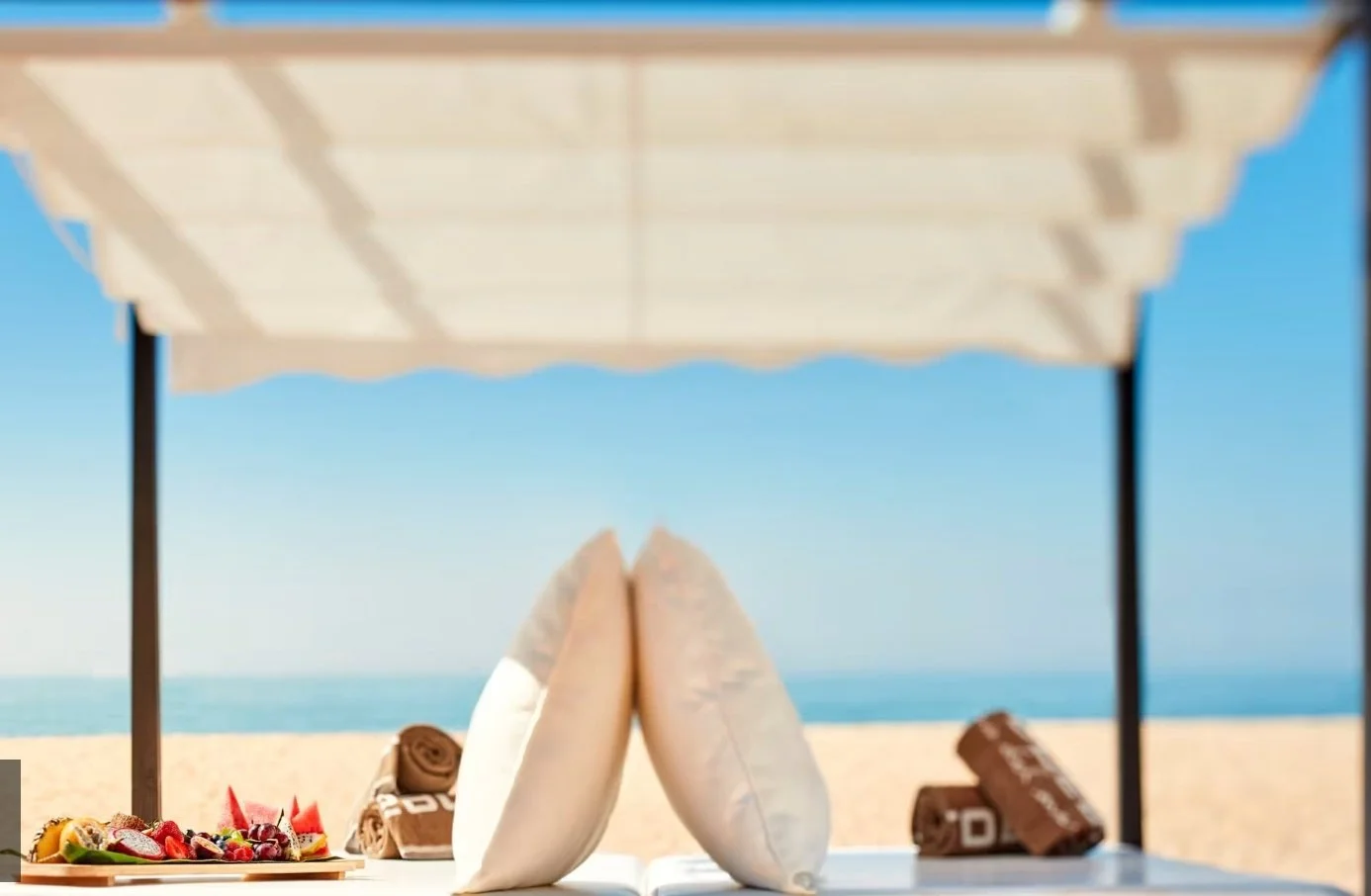 Beach scene with a canopy, two rolled towels, a plate of fresh fruit, and chocolate bars under the canopy, with sand and ocean in the background.