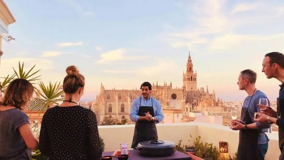 People gathered on a rooftop terrace with a city skyline featuring a historic cathedral in the background at sunset.