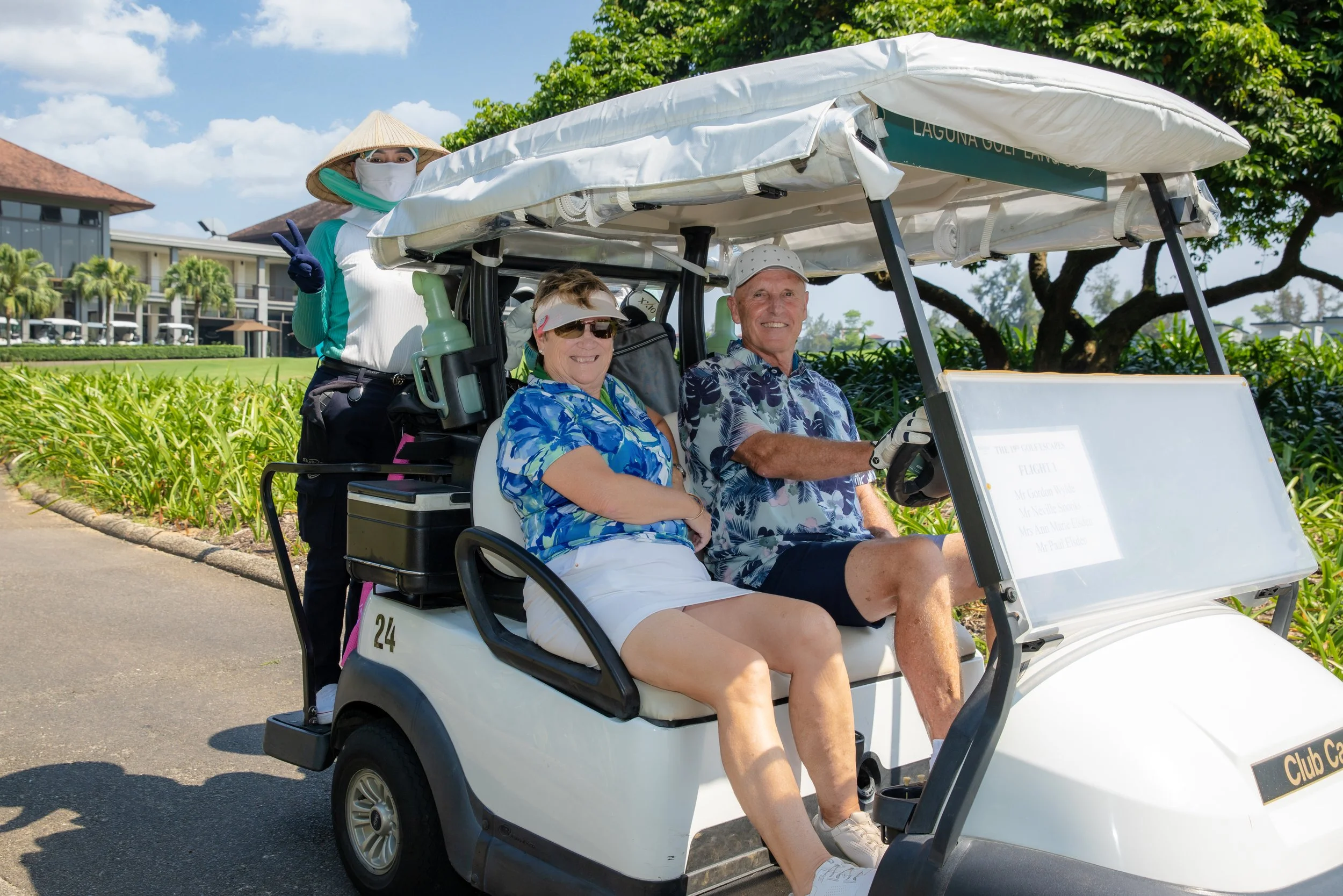Two seated older adults with light skin on a golf cart, surrounded by green trees and plants, with another person standing behind them making a peace sign.