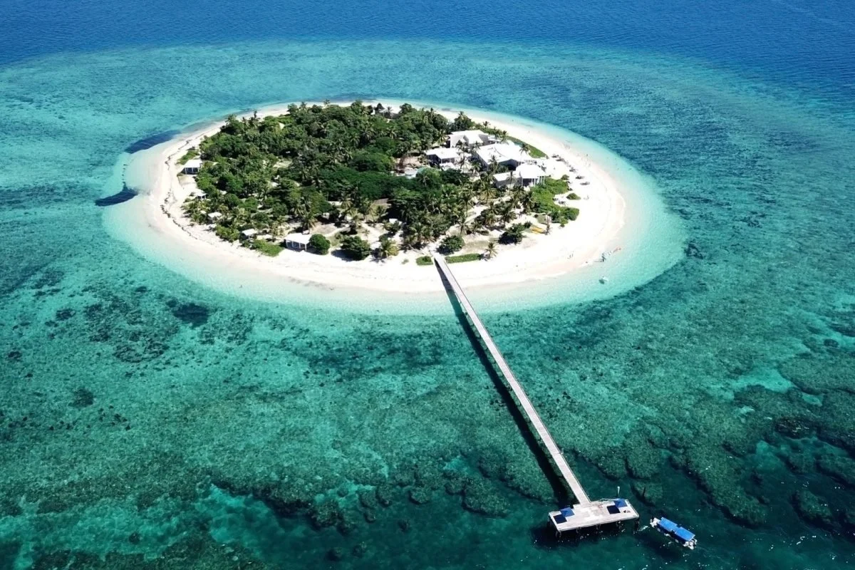Aerial view of a Malamala Island with a bridge extending to a floating dock, surrounded by clear turquoise water and coral reefs, which is a day trip activity on the Fiji hosted Golf Tour with The 19th Golf Escapes.