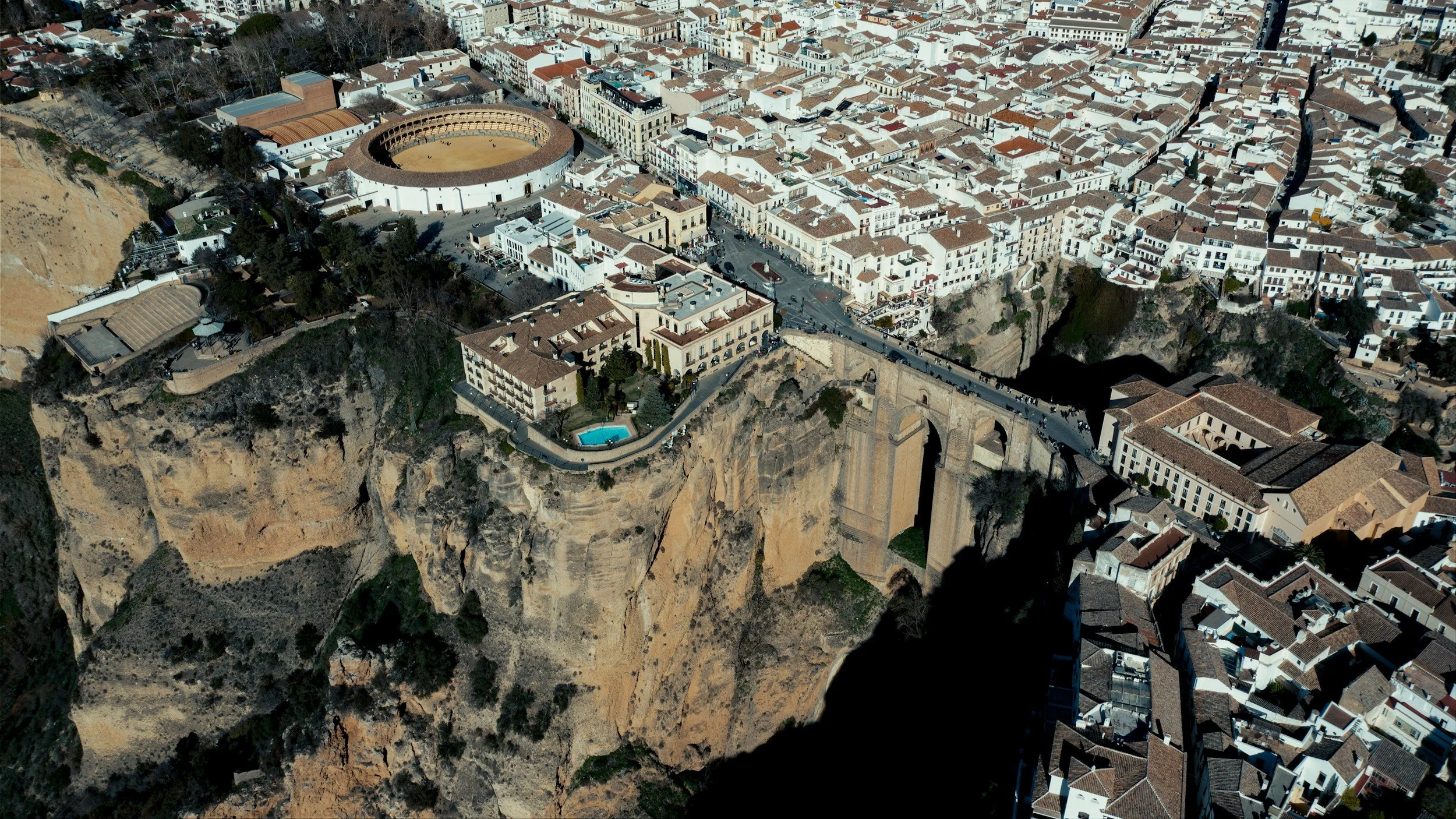 An aerial view of a city built on a steep cliff, featuring the famous bullring with a round shape and terracotta roof, surrounded by densely packed buildings with terracotta roofs, and a bridge connecting parts of the city over the canyon.