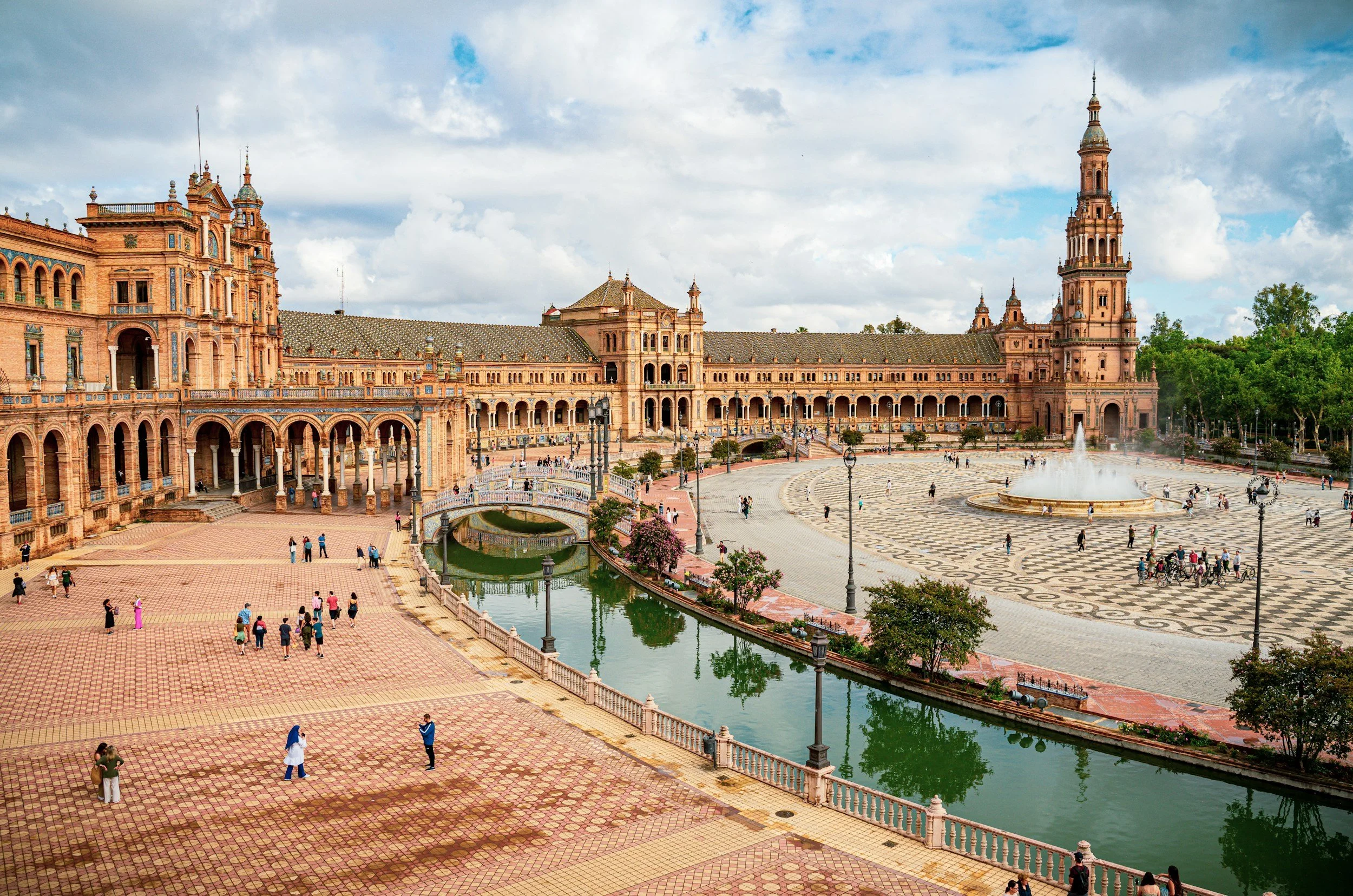 View of Plaza de España with its ornate pink and orange architecture, a central fountain, and a checkerboard pattern floor, with people walking and some riding bicycles on a partly cloudy day.