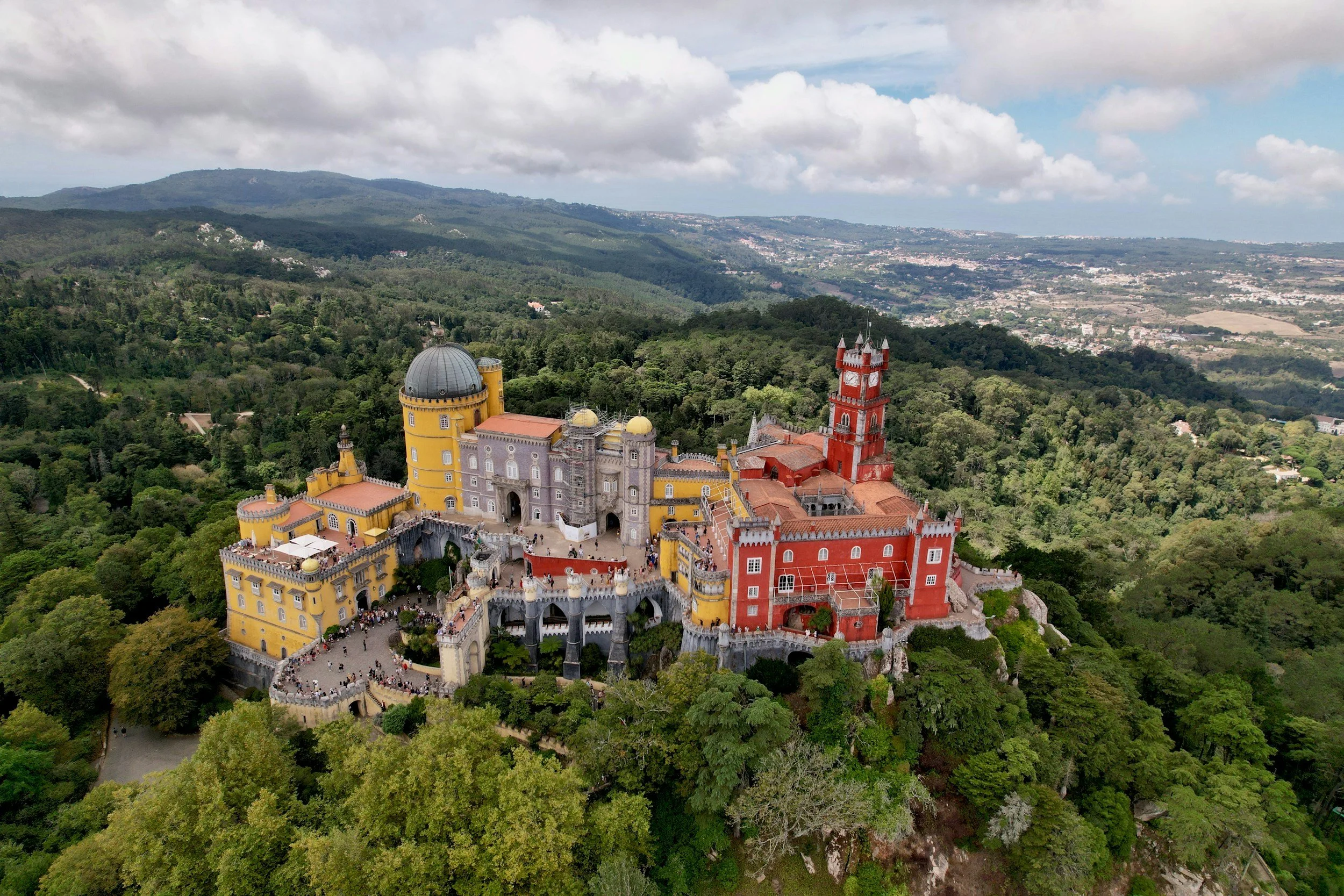 Aerial view of a colorful castle surrounded by lush green forest with rolling hills and cloudy sky in the background.