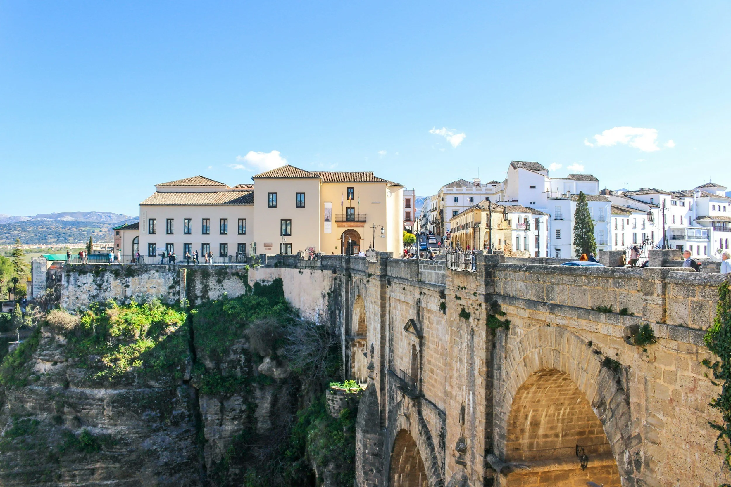 A scenic view of a European town with white buildings, a stone bridge, and a deep gorge with greenery below. The sky is clear and blue with a few clouds.