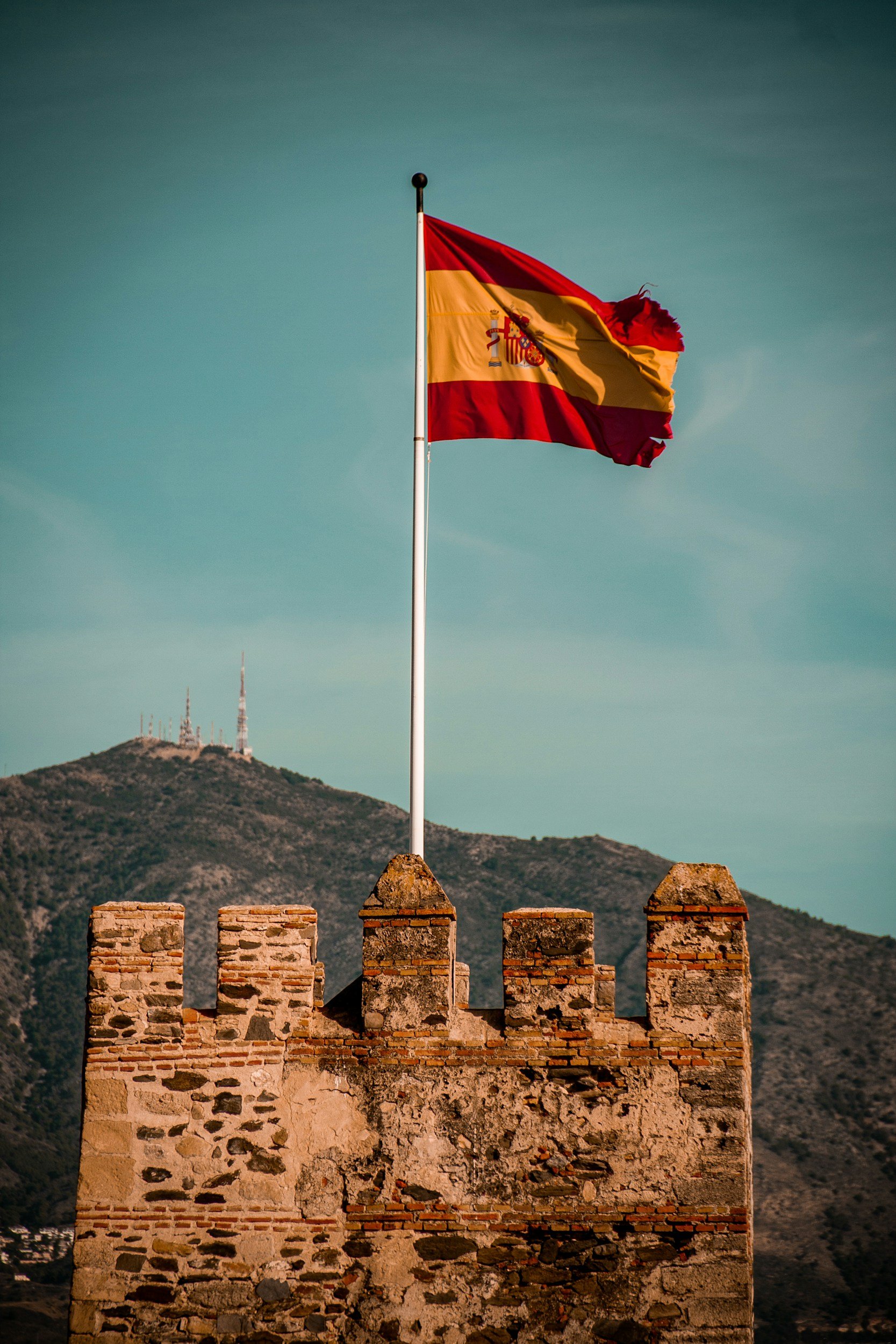 An American flag flying on a flagpole in front of a stone wall with a mountainous background and clear sky.