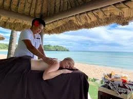 A woman receiving a massage on the beach under a thatched roof, with the ocean and sky in the background.
