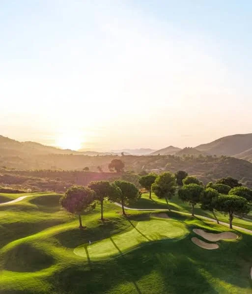 Sunset over a golf course with green fairways, sand bunkers, and scattered trees in a hilly landscape.