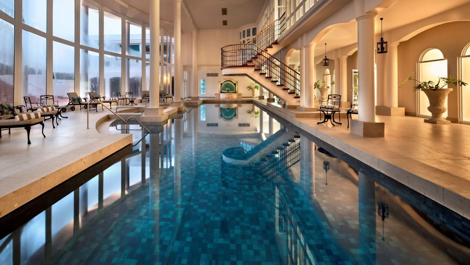 Indoor hotel swimming pool with lounge chairs, large windows, and elegant decor.