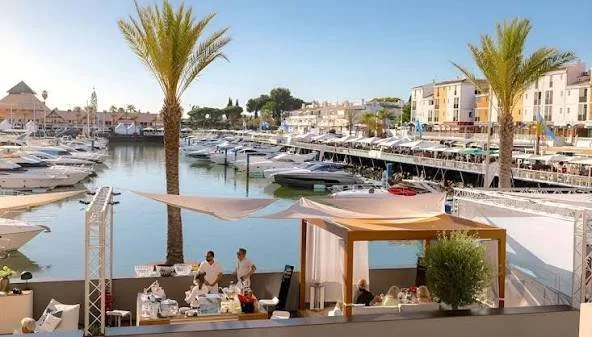 A marina with boats docked, palm trees, people dining at an outdoor waterfront restaurant, and buildings in the background.
