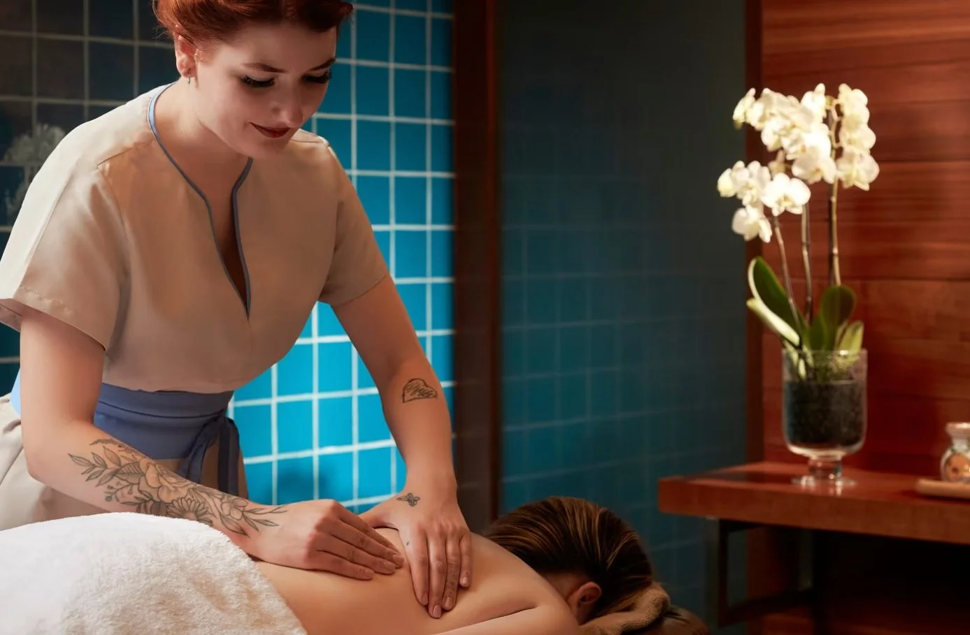A woman receiving a back massage in a spa room with blue tiled wall and a vase of white orchids.