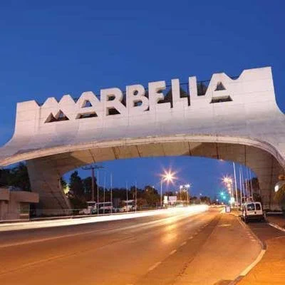 Marbella sign arch over a street at dusk with streetlights and cars.