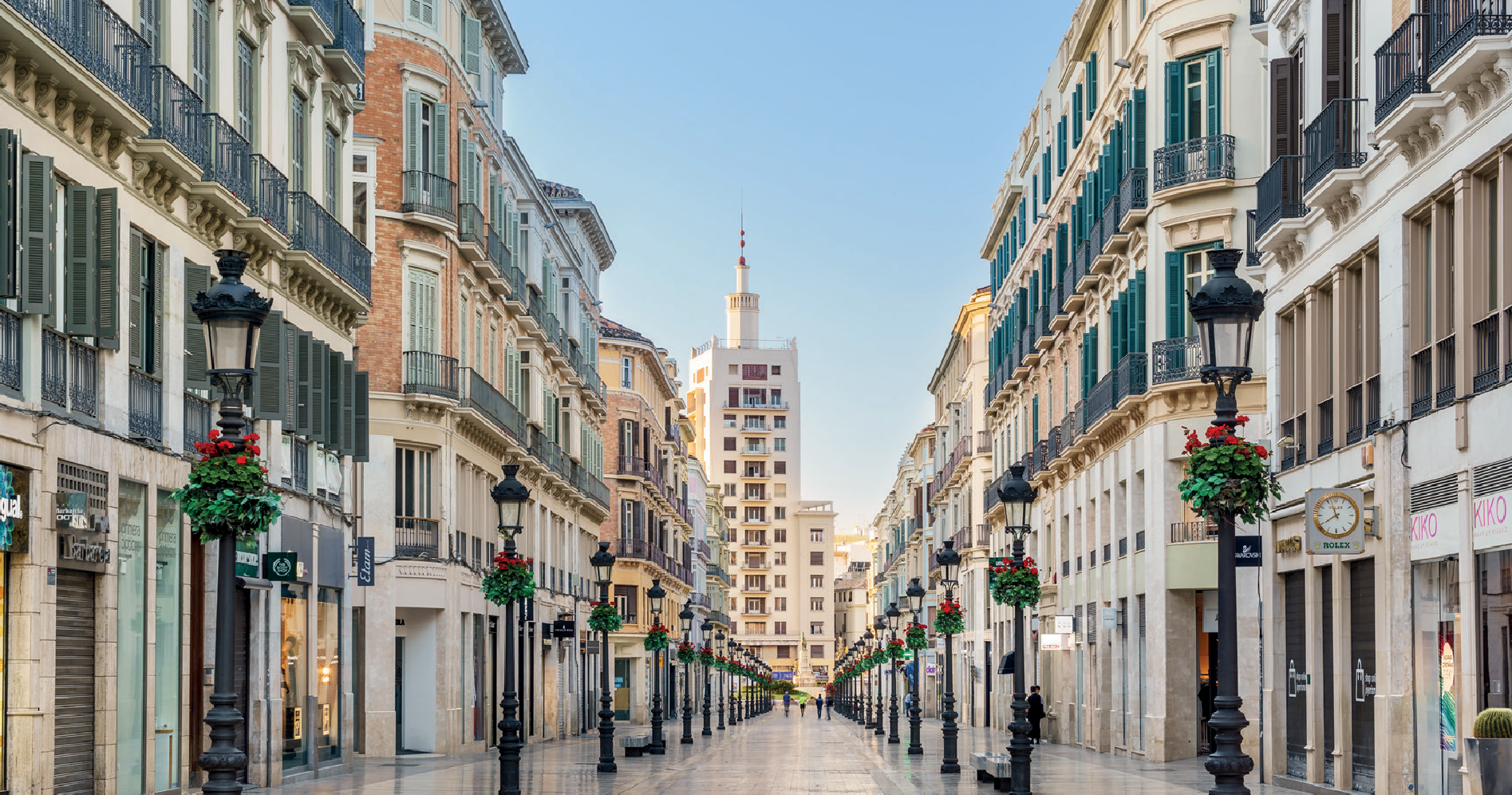 A wide pedestrian street lined with multi-story buildings on both sides, featuring balconies and large windows, under a clear blue sky. Black street lamps with hanging flower baskets are evenly spaced along the street. In the distance, there is a tall building with a spire.