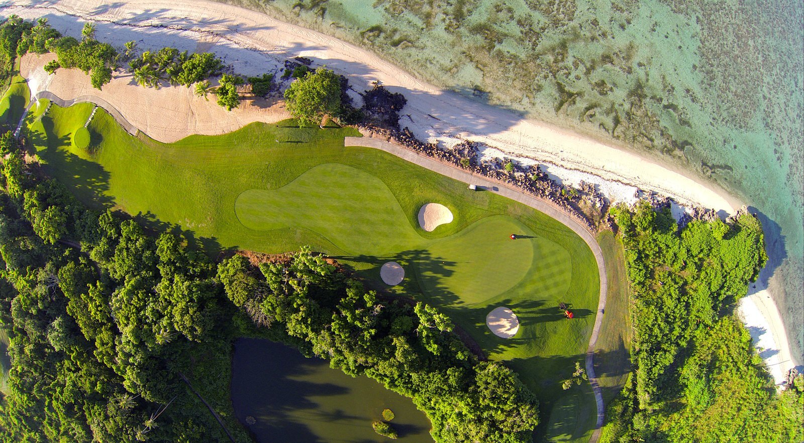 Aerial view of a golf course near a beach, showing green fairways, putting greens, sand bunkers, tall trees, and the ocean shoreline with clear water and coral reefs.