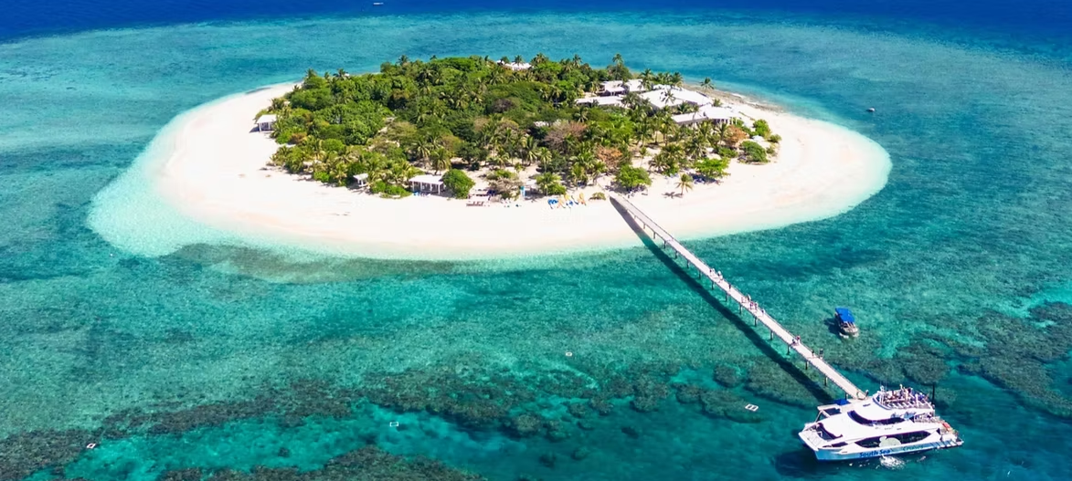 A tropical island with lush green trees and white sandy beaches surrounded by clear turquoise water, connected to a dock with boats.