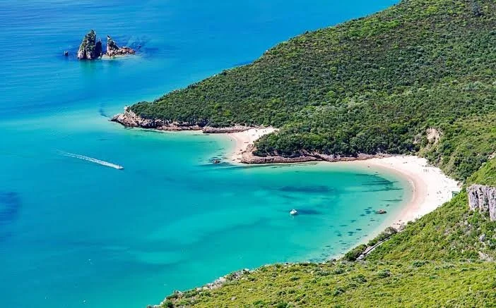 A scenic view of a turquoise bay with a small sandy beach, surrounded by green hills and a distant rock formation in the ocean.