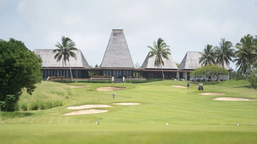 A golf course with sand bunkers, green grass, and palm trees in front of a large building with steep roofs, under a cloudy sky.