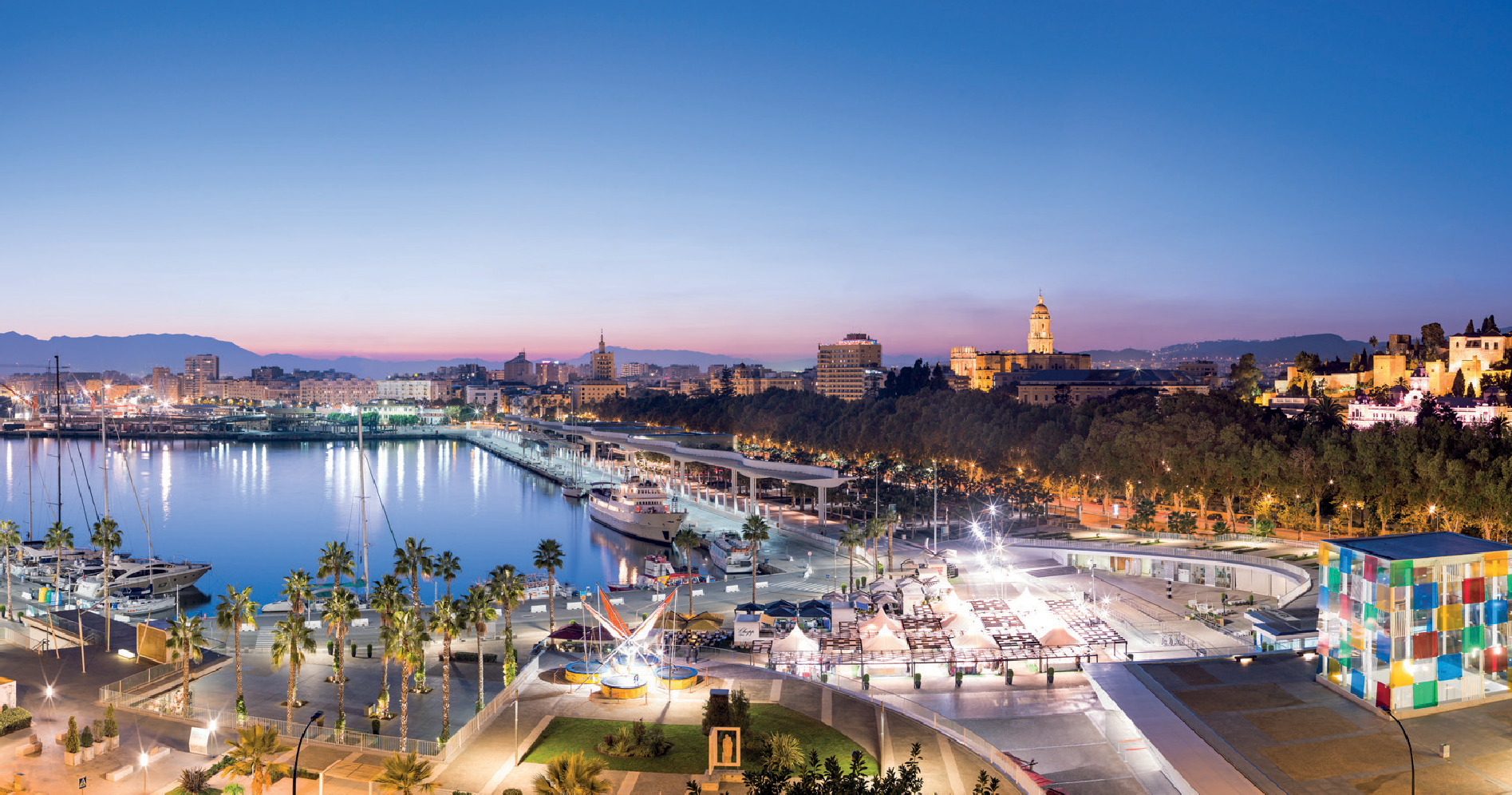 A city harbor at dusk with yachts docked in calm waters, surrounded by palm trees, with illuminated buildings and a colorful art installation in the foreground.