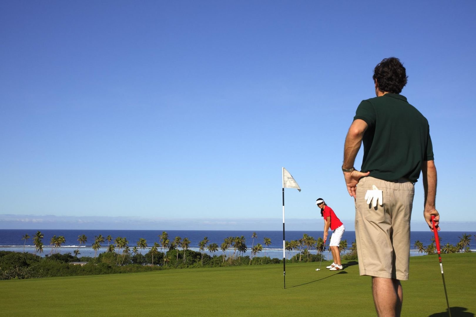 Two people playing golf on a course near the ocean, with palm trees and a blue sky in the background.