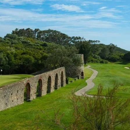 A golf course with a stone bridge or aqueduct, green fairway, and trees in the background under a blue sky with scattered clouds.