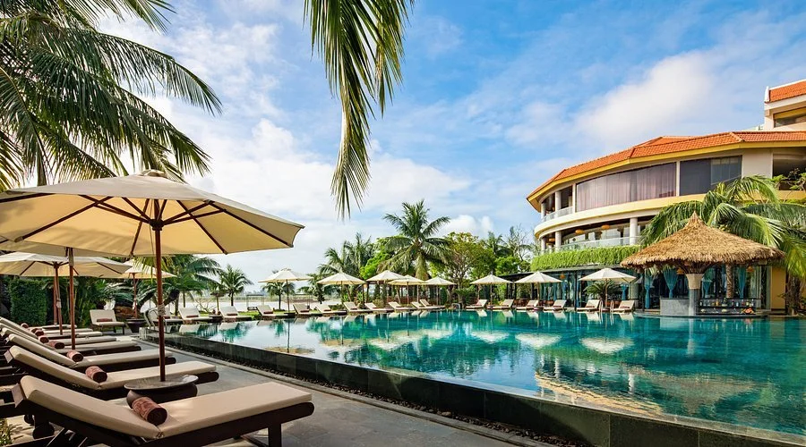 Resort pool area with lounge chairs, umbrellas, palm trees, and a large building in the background under a partly cloudy sky.