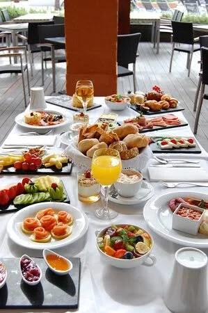 A breakfast spread on a table with various dishes, including pastries, fruit, cereal, juice, and eggs, set in a bright dining area with large windows.