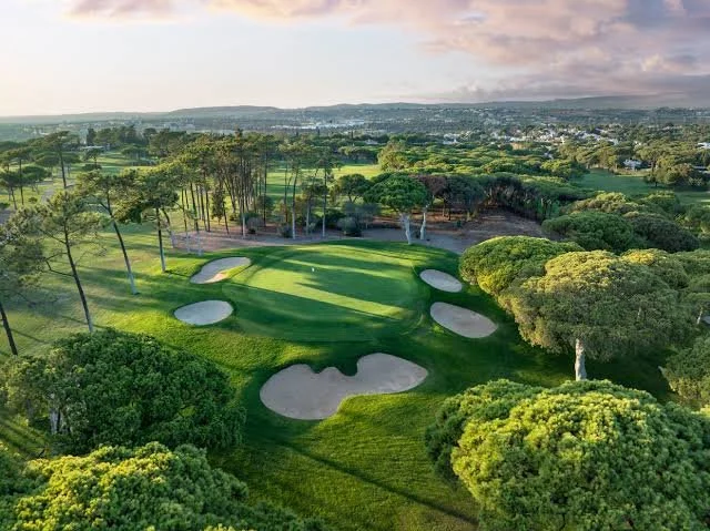 Aerial view of a golf course with sand traps, green fairways, trees, and a distant landscape under a cloudy sky.