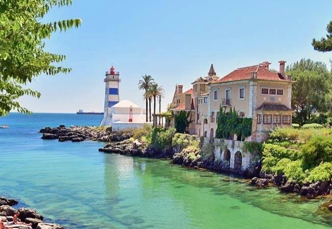 Coastal scene with historic houses, palm trees, lighthouse, and calm green water under a clear blue sky.