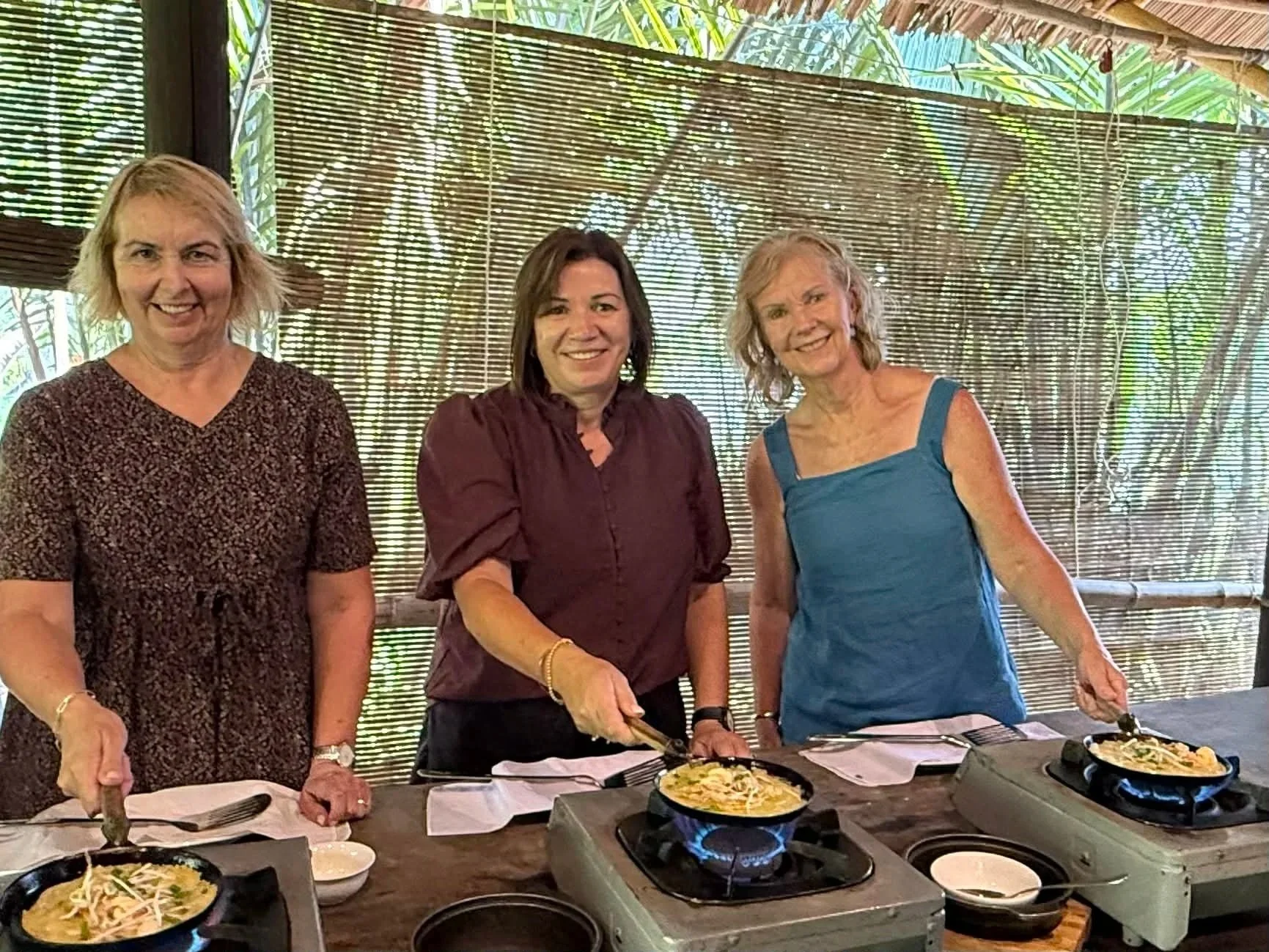 Three women cooking on portable stoves outdoors with a bamboo and greenery background.