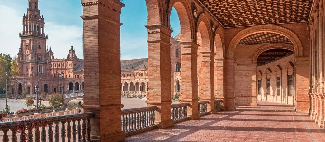 View of a historic building with a brick terrace, arched columns, and a tall clock tower in the background, under a clear blue sky.