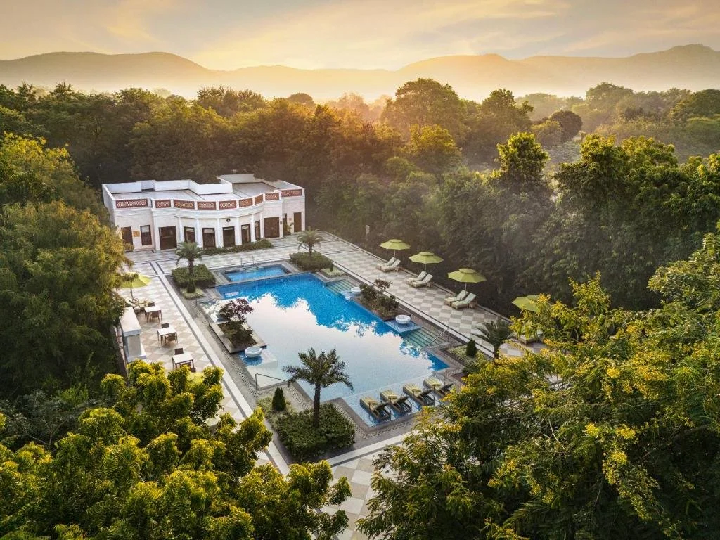 An aerial view of a luxurious outdoor swimming pool surrounded by lush green trees and a scenic mountain landscape at sunset.