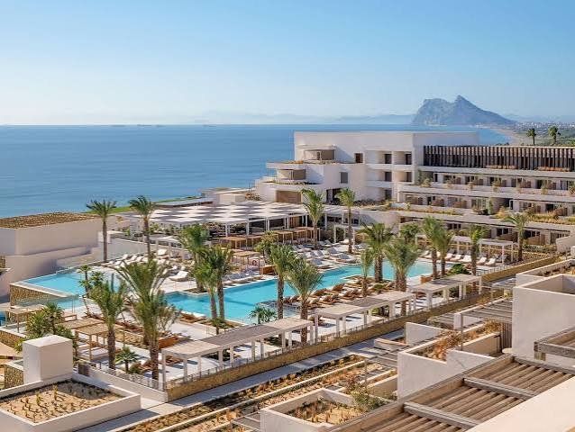 Luxury beach resort with a large swimming pool surrounded by palm trees, lounge chairs, and white buildings overlooking the ocean with a clear blue sky.