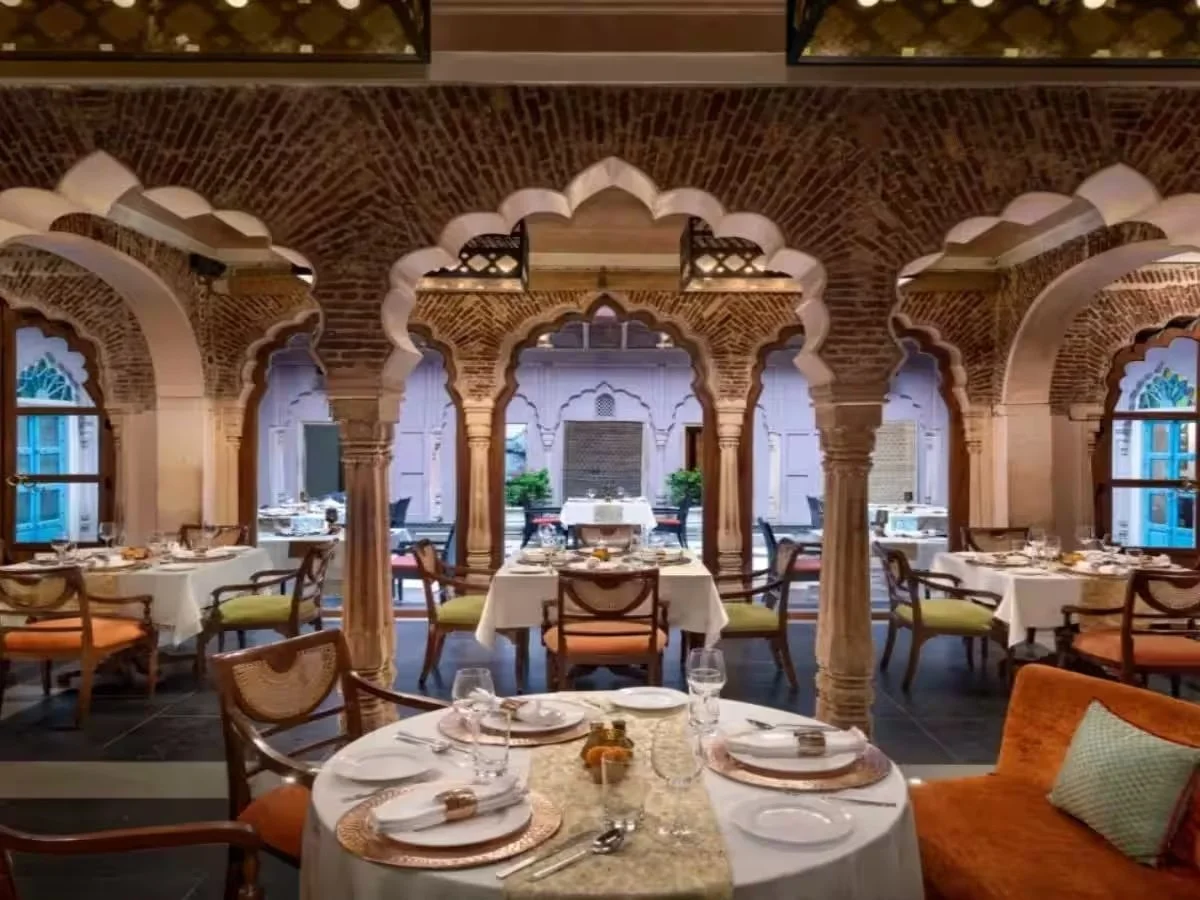 Elegant restaurant dining room with arched brick architecture, tables set with white tablecloths, plates, silverware, and glasses, and chairs with colorful cushions, overlooking a courtyard with outdoor seating.