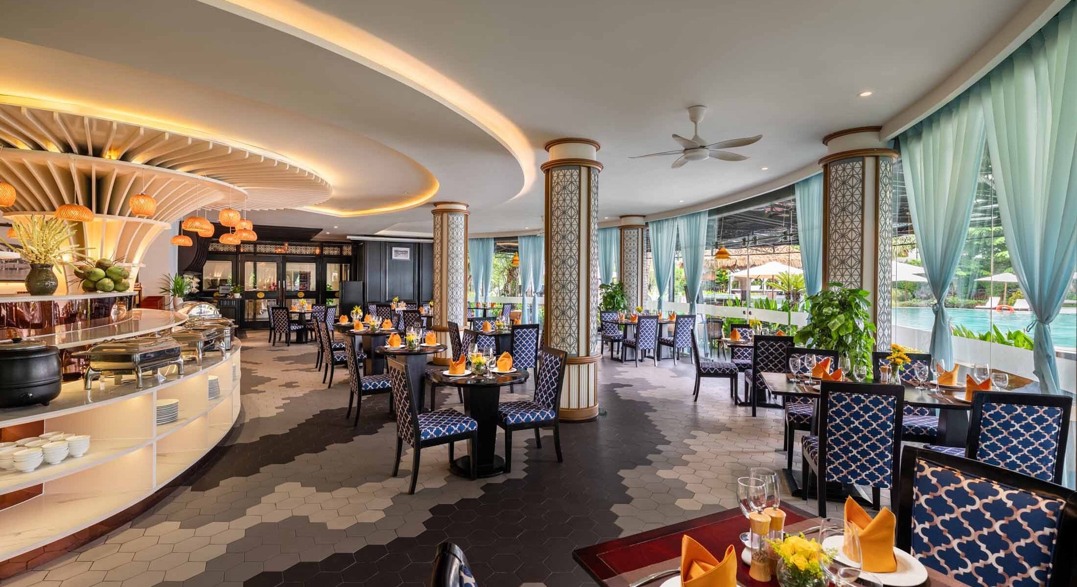 Elegant restaurant dining area with tables set for service, decorated with orange napkins and small flower arrangements. There are large windows with blue curtains allowing natural light and a view of a pool outside. The interior features stylish columns, potted plants, and a curved ceiling with warm lighting.