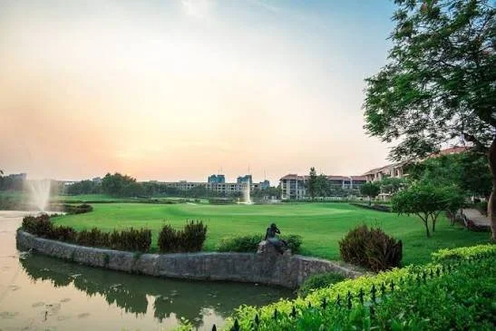 A park at sunset with a water fountain, lush green grass, bushes, and trees, with buildings in the background.