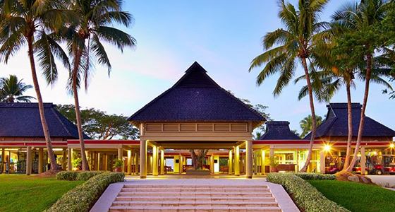 A tropical resort entrance with palm trees, a paved walkway, and a traditional-style building.