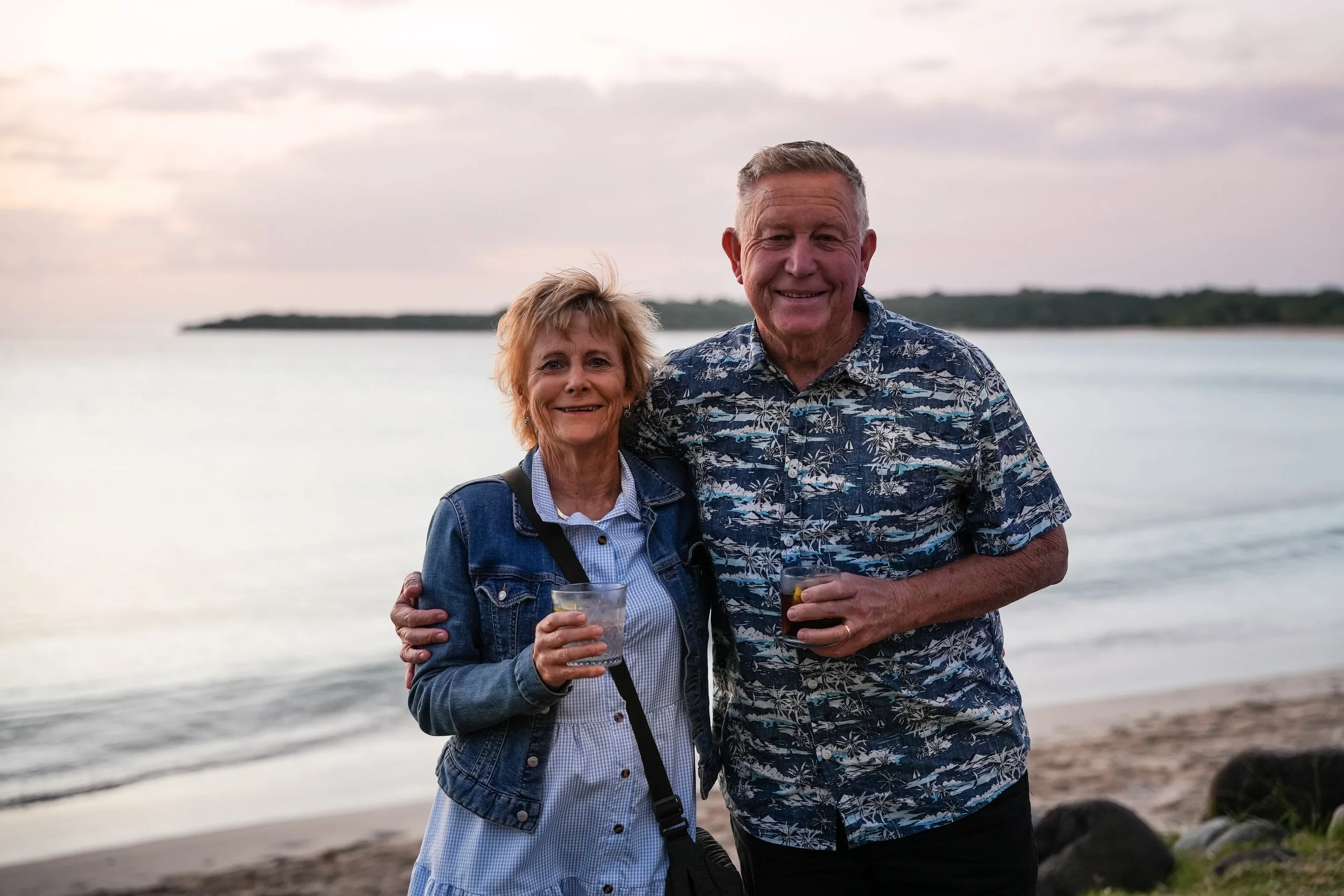 An elderly man and woman smiling and holding drinks on a beach, with water and a distant coastline in the background during an overcast evening.