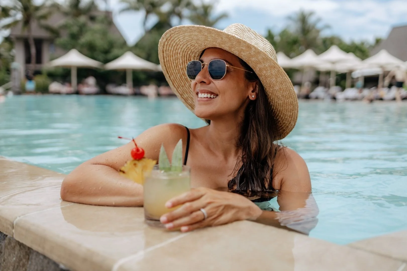Woman relaxing in a swimming pool with a straw hat and sunglasses, holding a tropical cocktail with a cherry garnish.