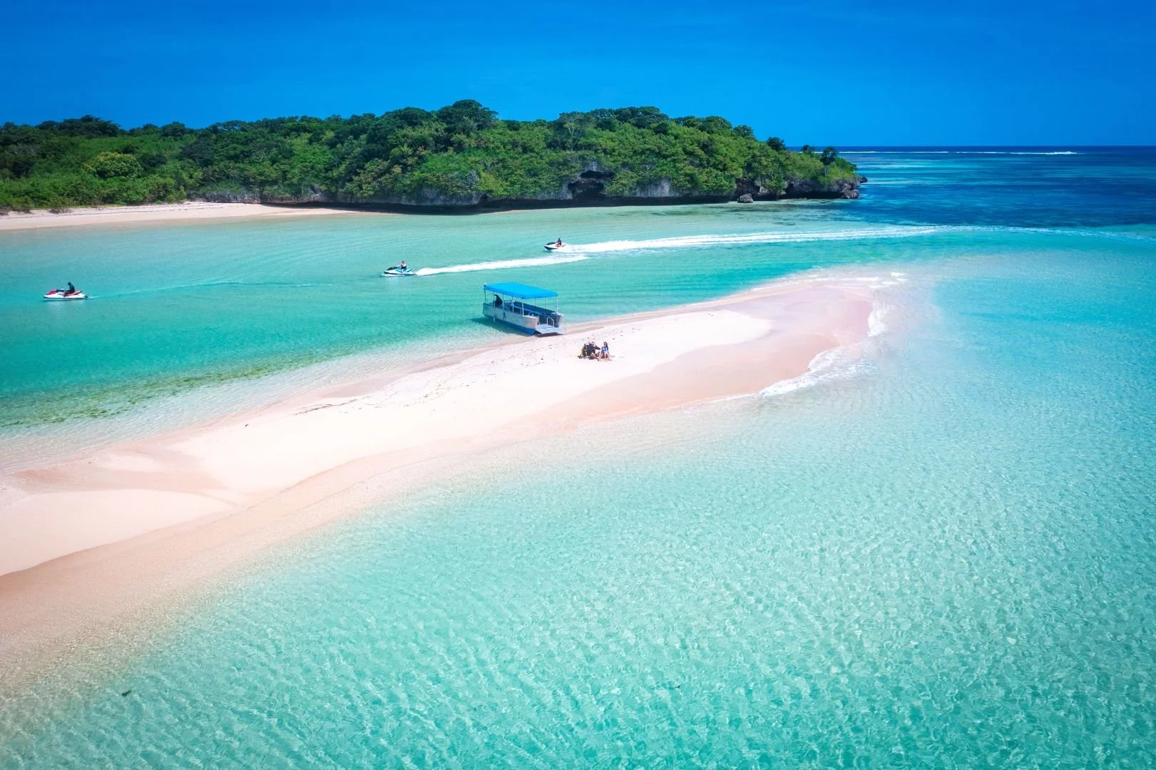 Tropical beach with clear turquoise water, a small sandy island, a boat, and three jet skis in the water. Dense green trees cover the background.