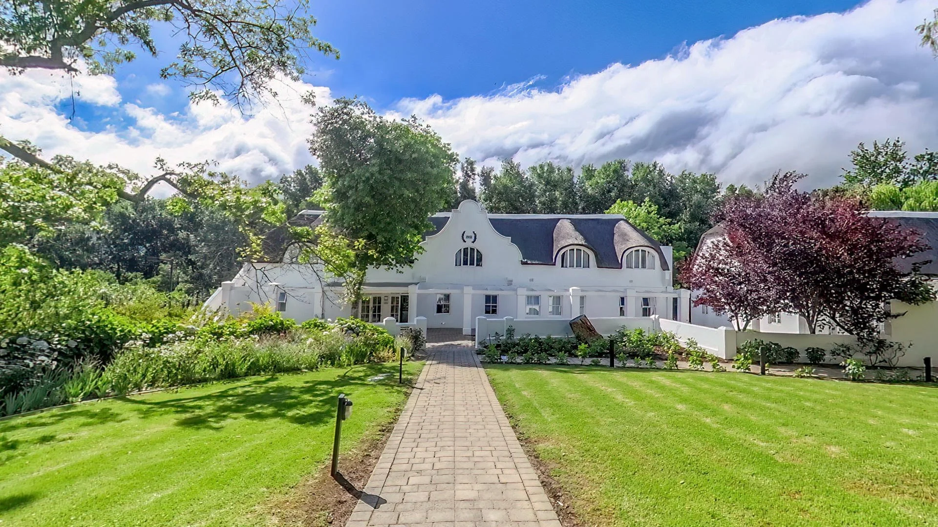 White house with a curved roof and multiple windows, surrounded by lush green lawns and trees, under a partly cloudy sky.