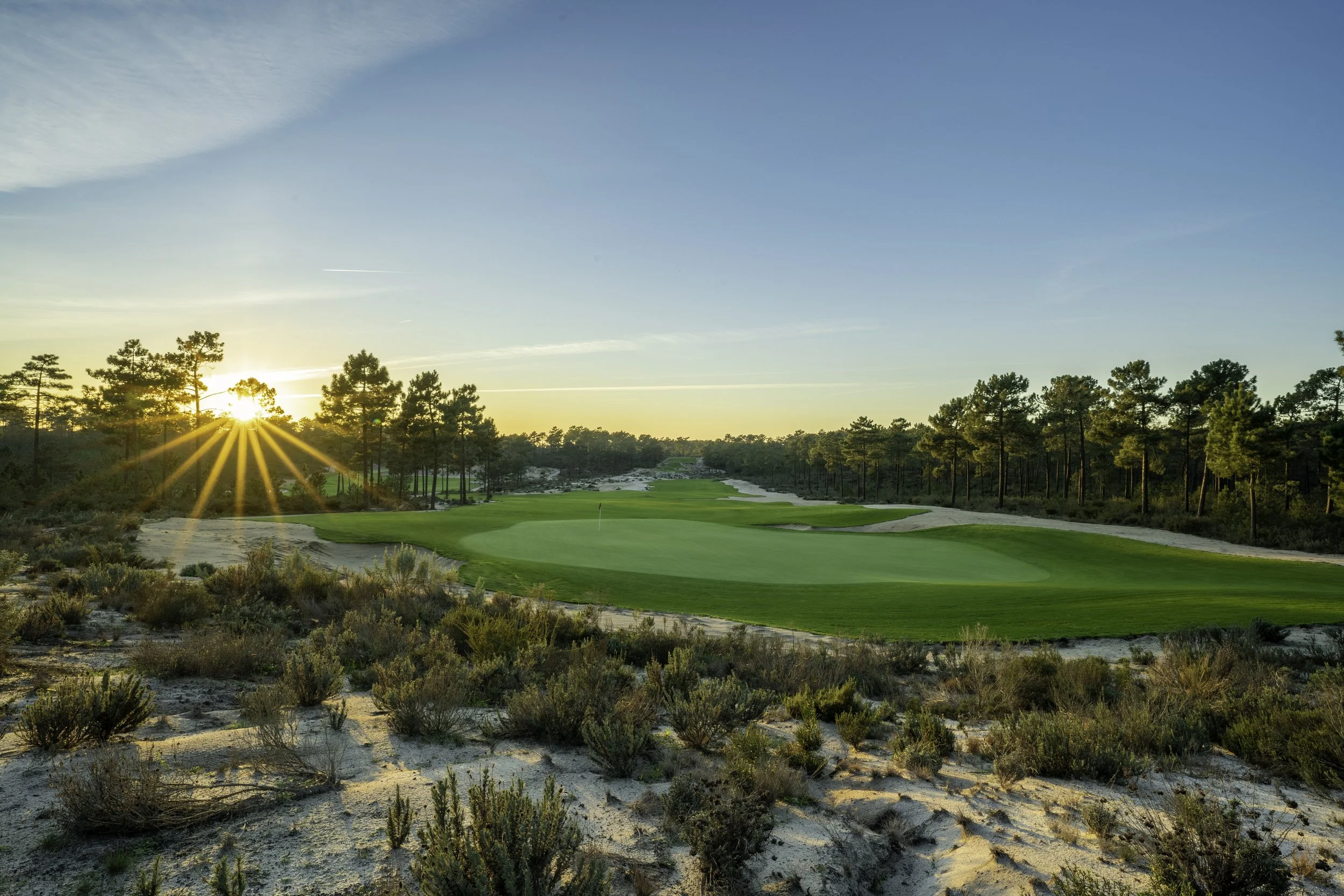 A golf course at sunrise with green fairways, surrounded by trees and sandy areas, under a partly cloudy blue sky.